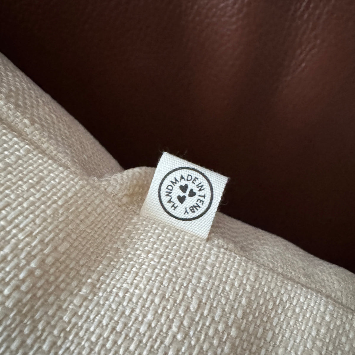 Close-up of a cream-colored woven fabric item with a small white sewn label that reads 'HANDMADE IN TENBYY' with a heart logo, set against a dark brown textured background.