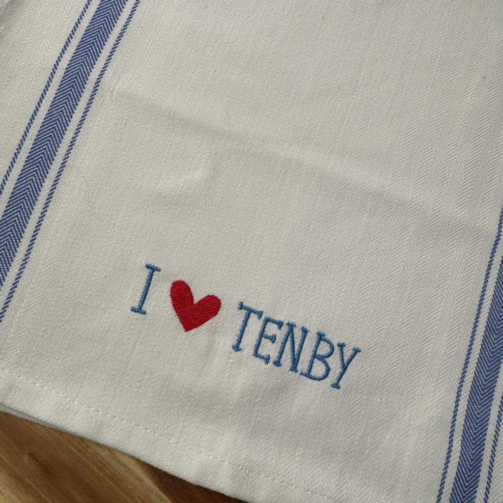 A white cotton kitchen towel with blue stripes on the sides and embroidered blue text reading 'I' followed by a red heart and 'TENBY' on a wooden surface.