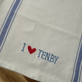 A white cotton kitchen towel with blue stripes on the sides and embroidered blue text reading 'I' followed by a red heart and 'TENBY' on a wooden surface.