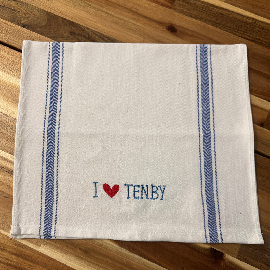 A white cotton tea towel with blue stripes and 'I [heart] TENBY' embroidered in blue and red, laid flat on a wooden surface.