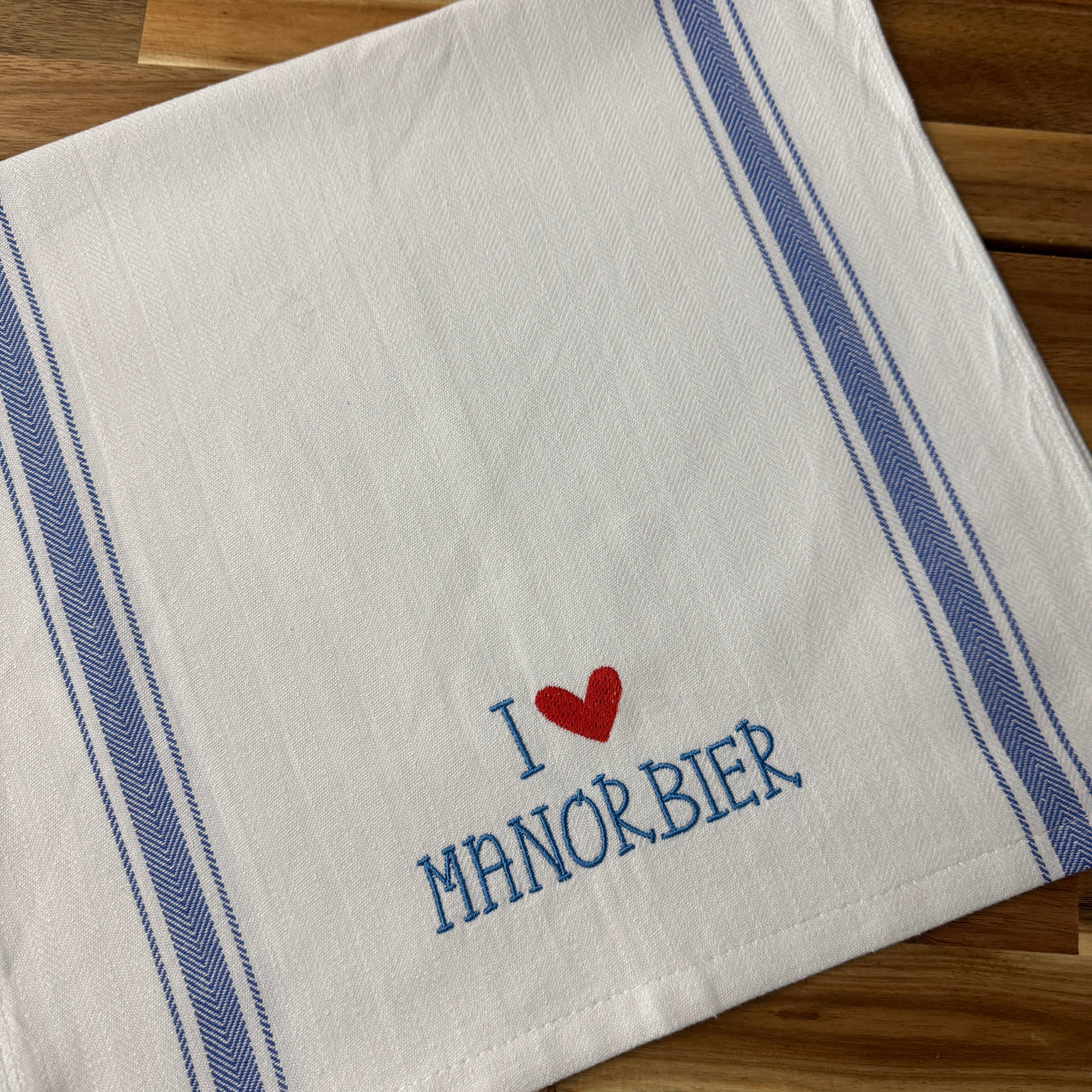 A white cotton kitchen towel with blue stripes and embroidered text reading 'I [red heart] MANORBIER' in blue, laid flat on a wooden surface.