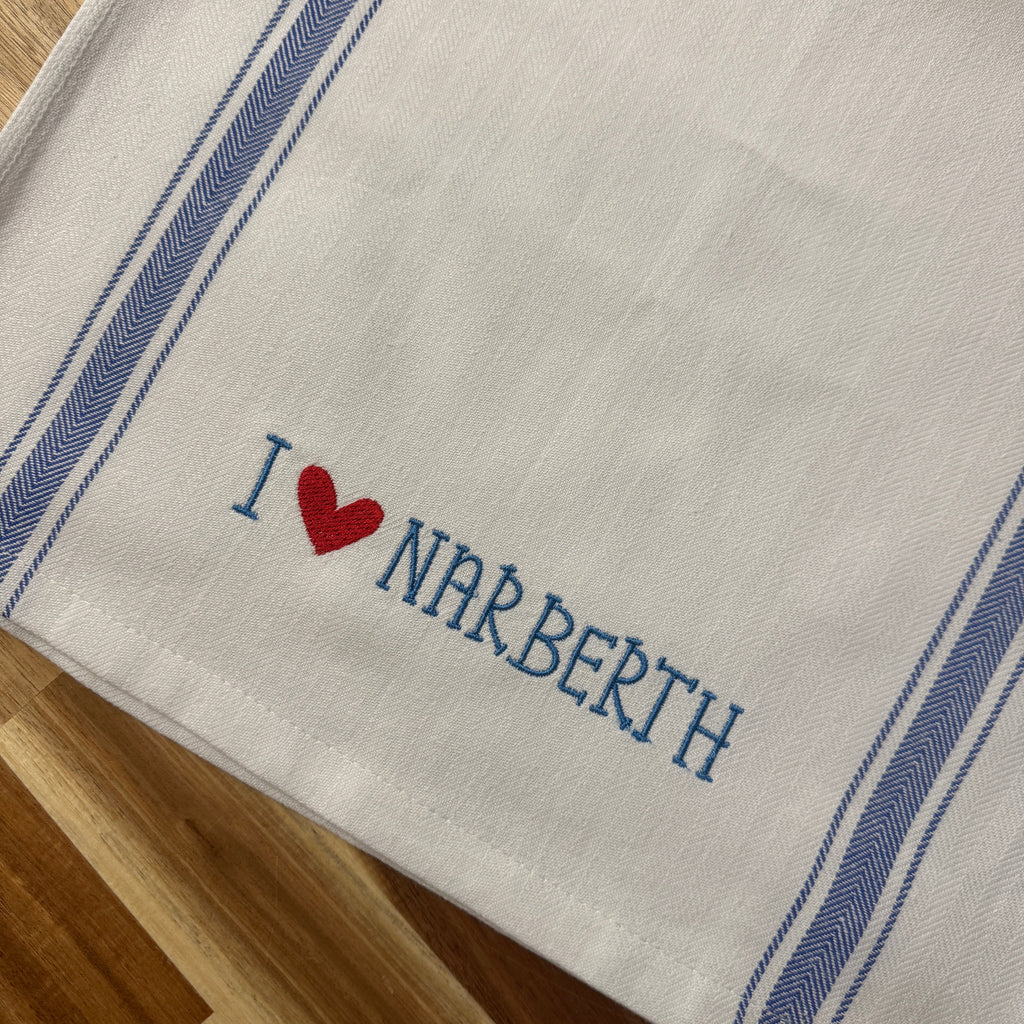 A close-up of a white woven textile, likely a towel or napkin, featuring blue striped borders and the embroidered text 'I [red heart] NARBERTH' in blue and red. The textile is laid flat on a wooden surface.