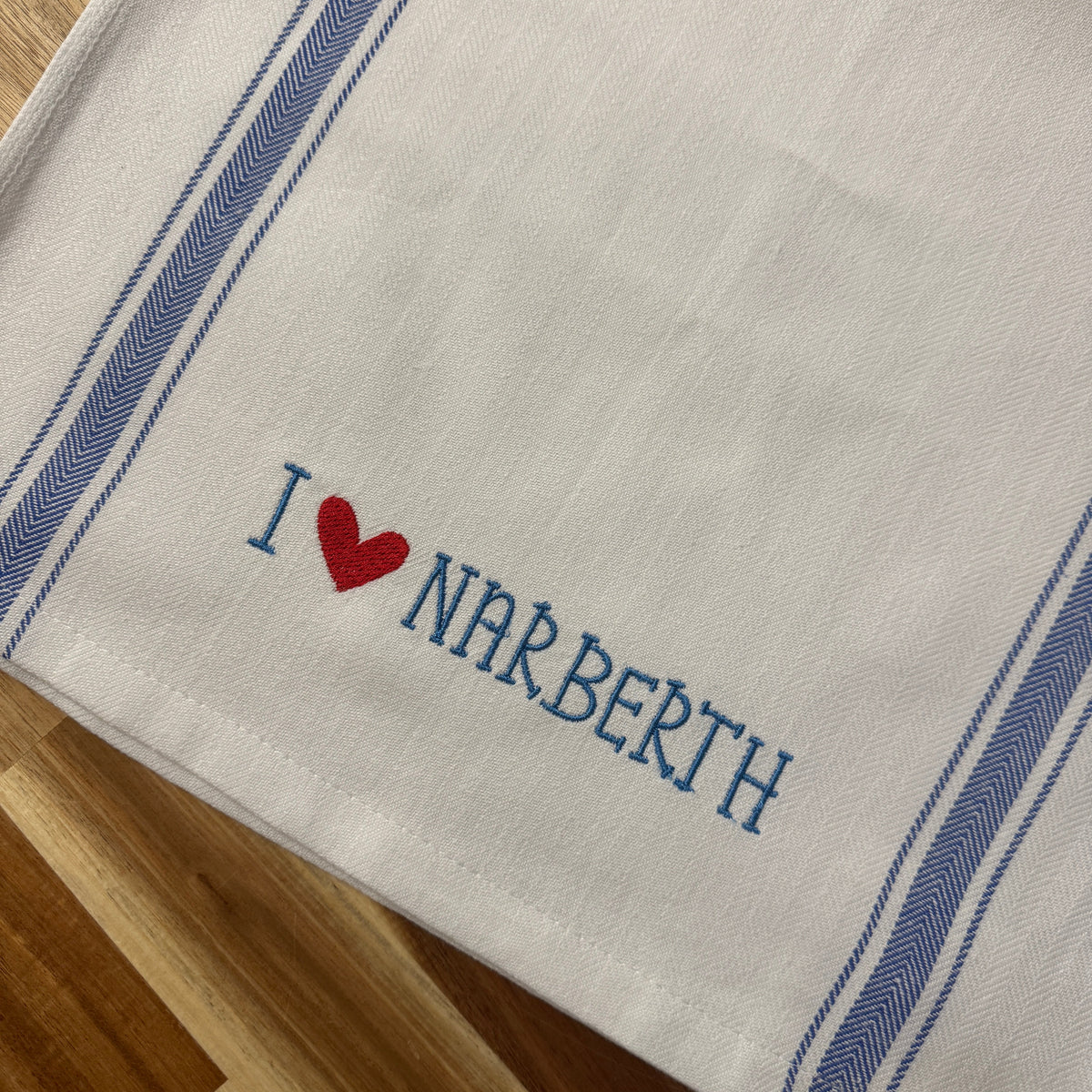 A close-up of a white woven textile, likely a towel or napkin, featuring blue striped borders and the embroidered text 'I [red heart] NARBERTH' in blue and red. The textile is laid flat on a wooden surface.