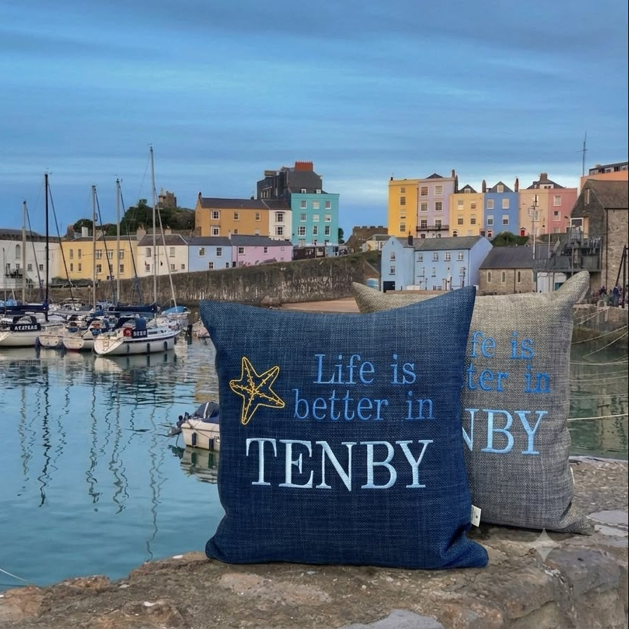 Two decorative throw pillows displayed on a stone pier in front of a harbor scene with boats and colorful buildings. The front pillow is navy blue with white text reading 'Life is better in TENBY' and a yellow starfish graphic, while the rear pillow is heather gray with matching blue text.