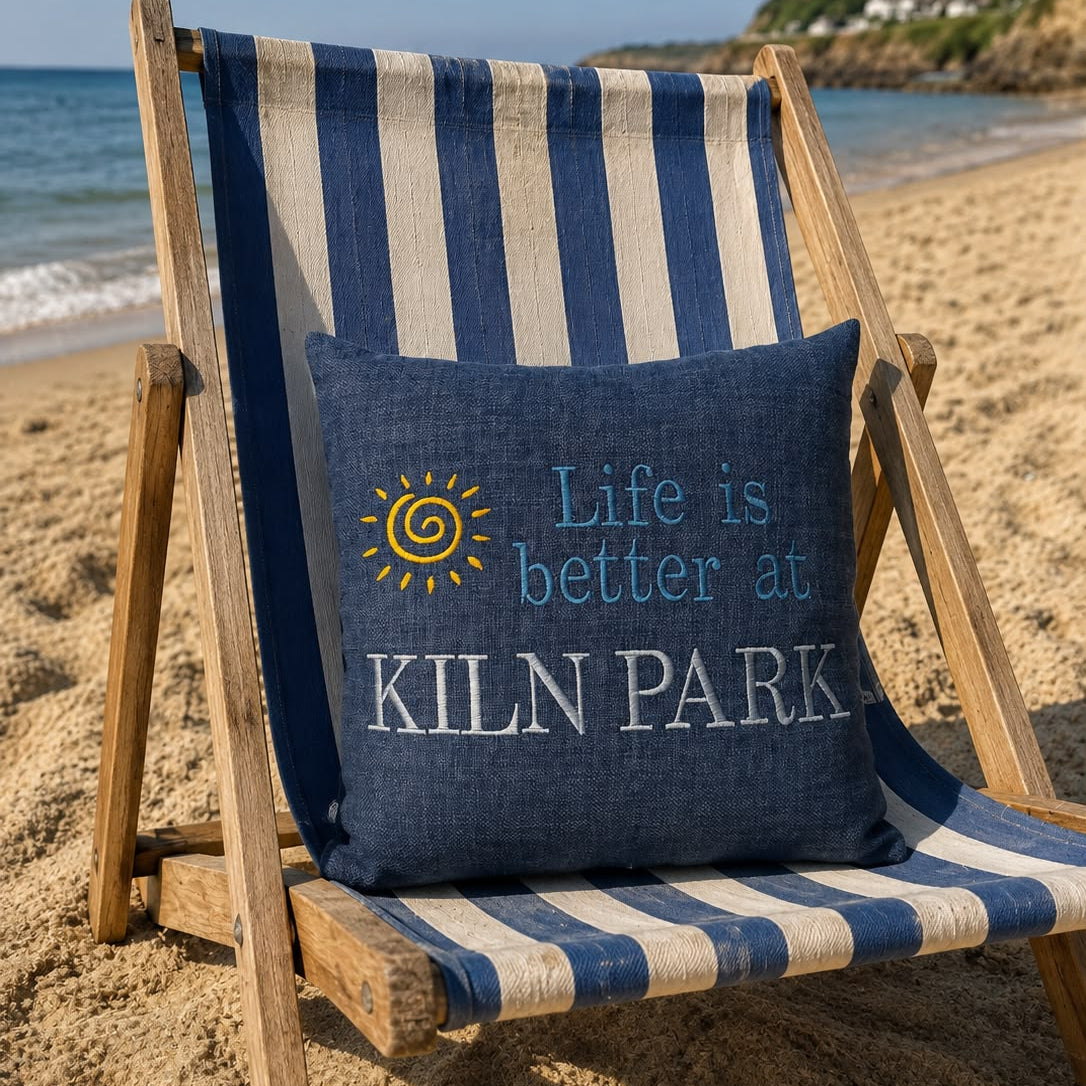 A navy blue decorative throw pillow with the text 'Life is better at KILN PARK' and a yellow sun graphic, resting on a blue and cream striped wooden beach chair on a sandy beach.