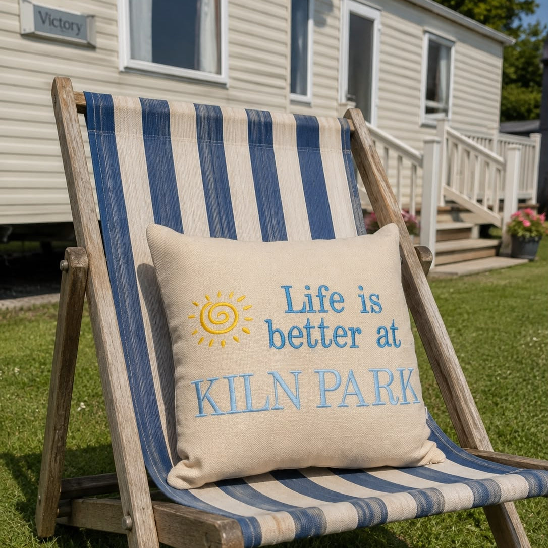 A beige throw pillow with blue and yellow embroidered text reading 'Life is better at KILN PARK' sits on a blue and beige striped wooden deck chair on a grassy lawn in front of a mobile home.