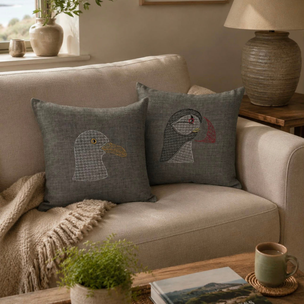 Two square gray throw pillows with embroidered puffin designs rest on a beige sofa in a styled living room setting.