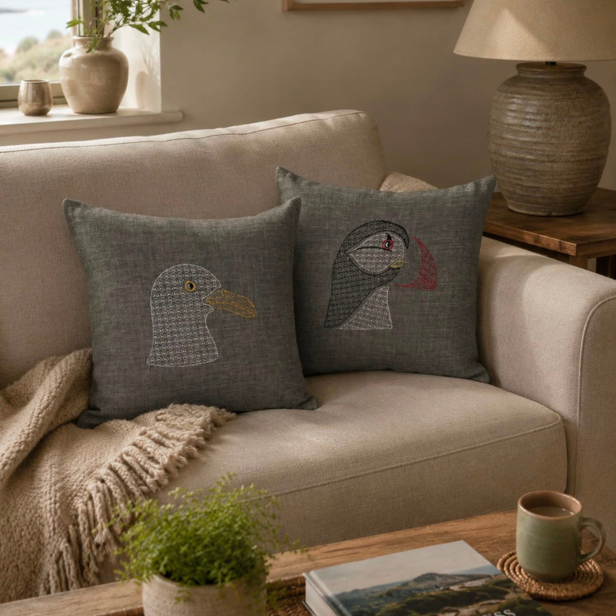 Two square gray throw pillows with embroidered puffin designs rest on a beige sofa in a styled living room setting.