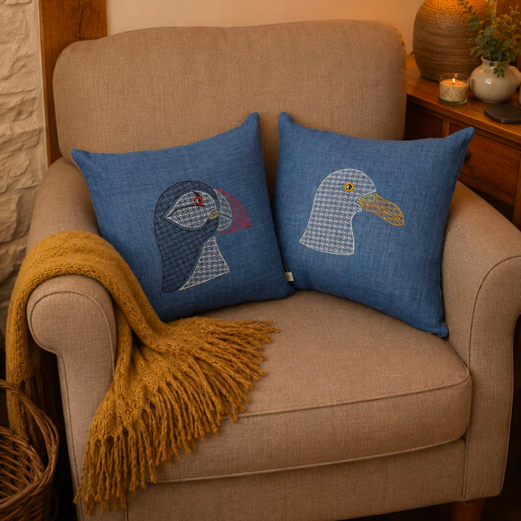 Two blue throw pillows with embroidered bird designs resting on a beige armchair in a cozy, warmly lit room. A mustard yellow throw blanket is draped over the arm of the chair.
