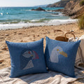 Two blue decorative throw pillows with patchwork puffin designs, placed on a beach blanket on sandy shore with the ocean and cliffs in the background.