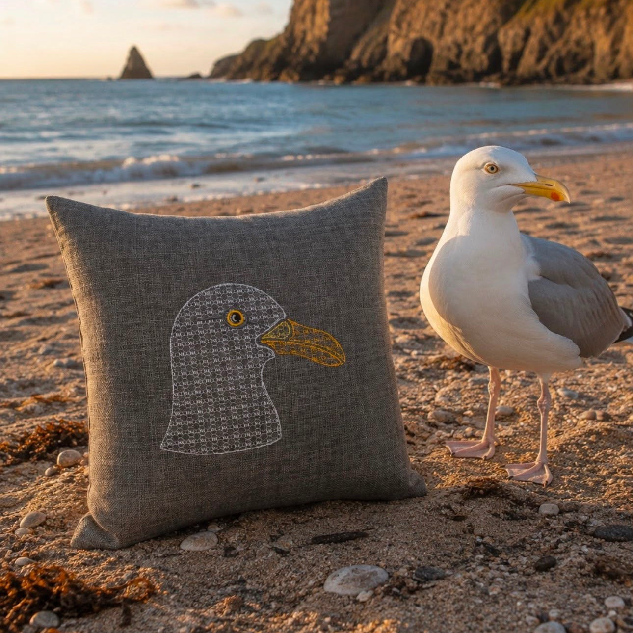 A square linen throw pillow with an embroidered seagull design sits on a sandy beach next to a real seagull, with ocean waves and rocky cliffs in the background.