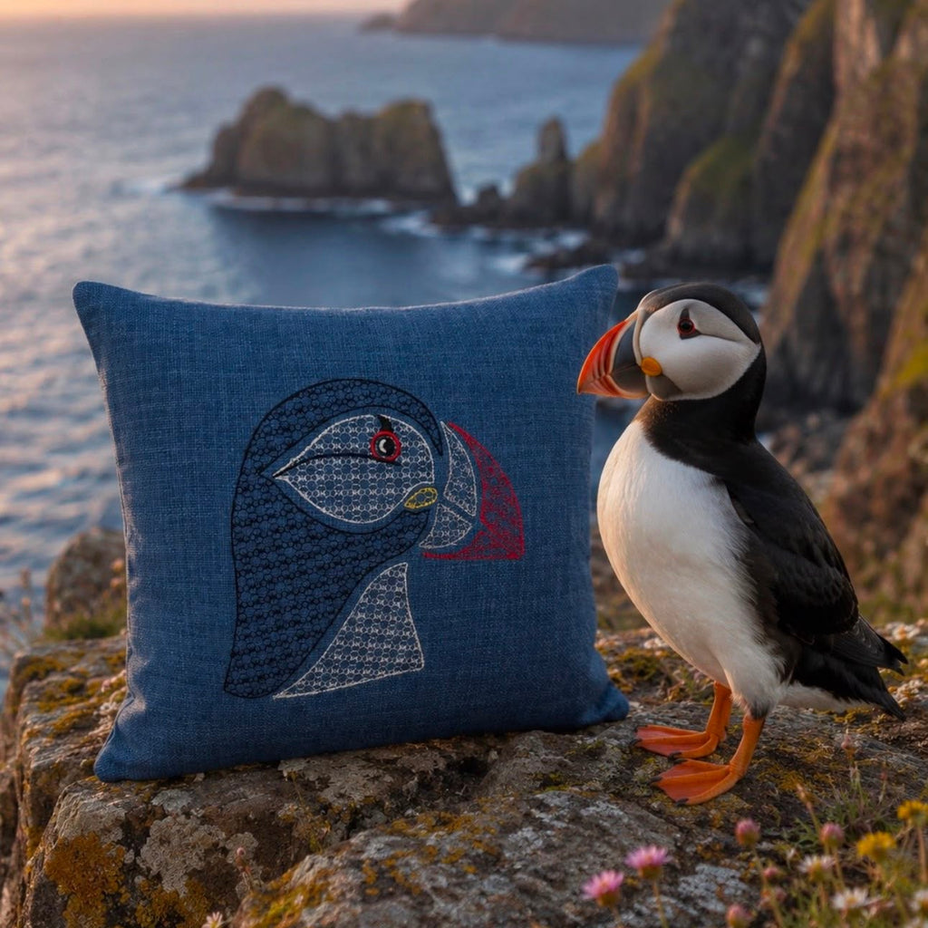 A square blue throw pillow with an embroidered puffin design sits on a rocky cliffside next to a real puffin bird, with the ocean and cliffs in the background at sunset.