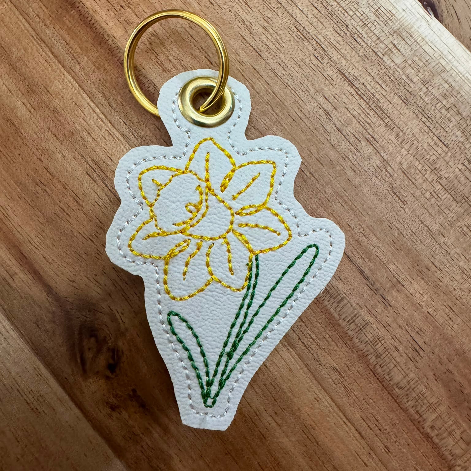 A white embroidered keychain charm shaped like a daffodil flower, featuring yellow petals, green leaves, and a gold metal key ring, shown on a wooden surface.