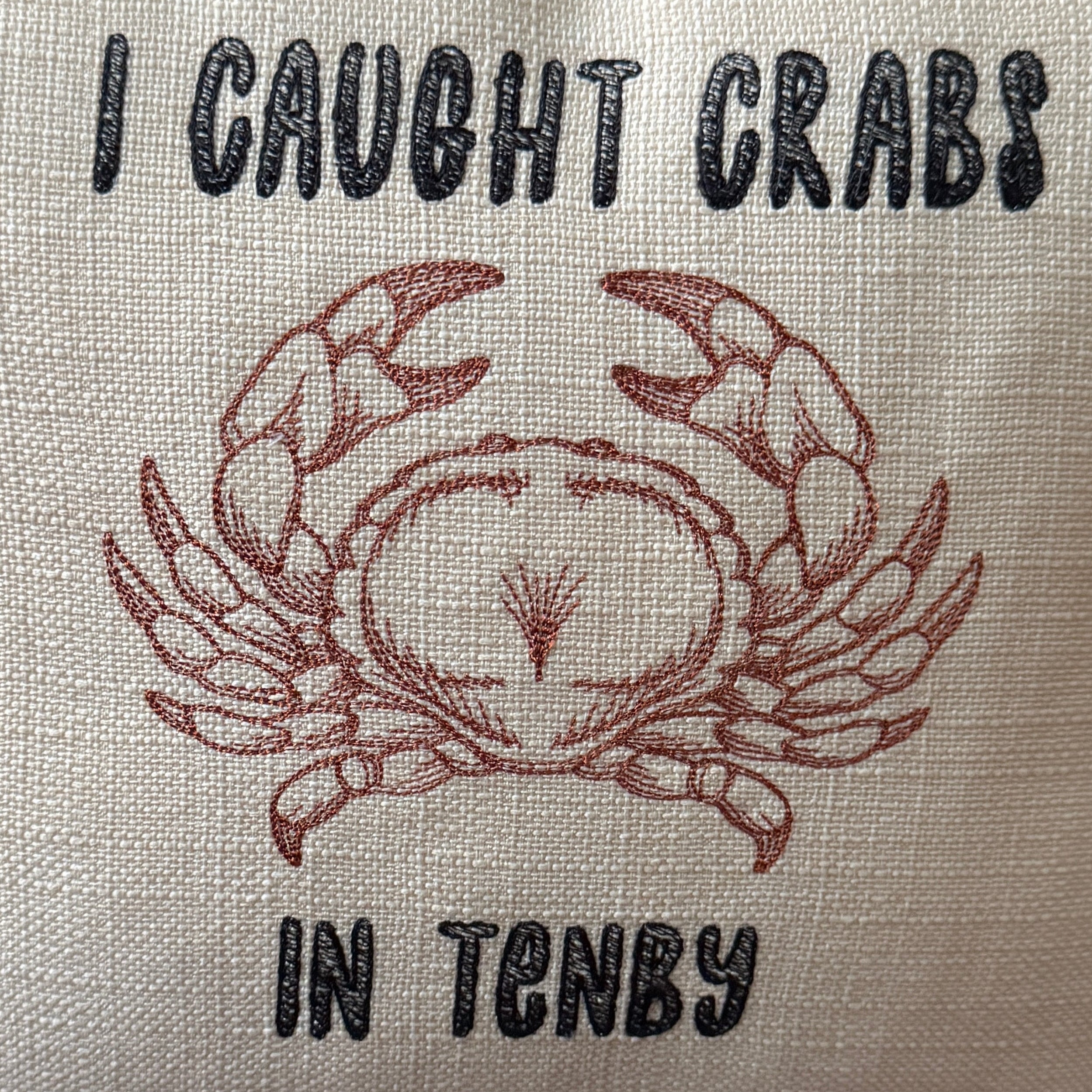 Close up of a square cream-colored  throw pillow with a humorous crab graphic and the text 'I CAUGHT CRABS IN TENBY' in black and reddish-brown embroidery, resting on a brown leather couch.