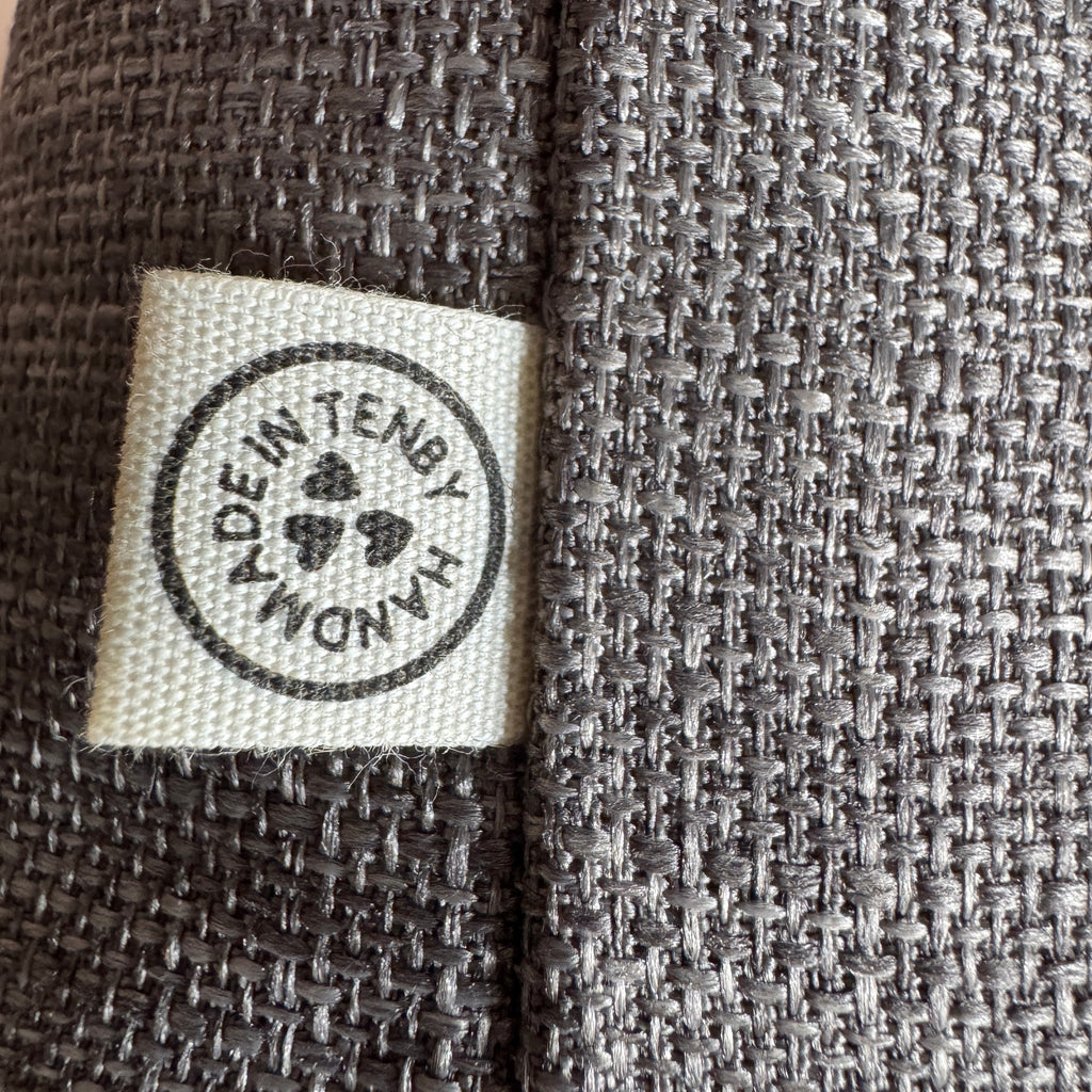 Close-up of a dark gray woven fabric with a square cream fabric label sewn onto the side. The label has a black circular stamp that reads 'MADE IN TENBY.