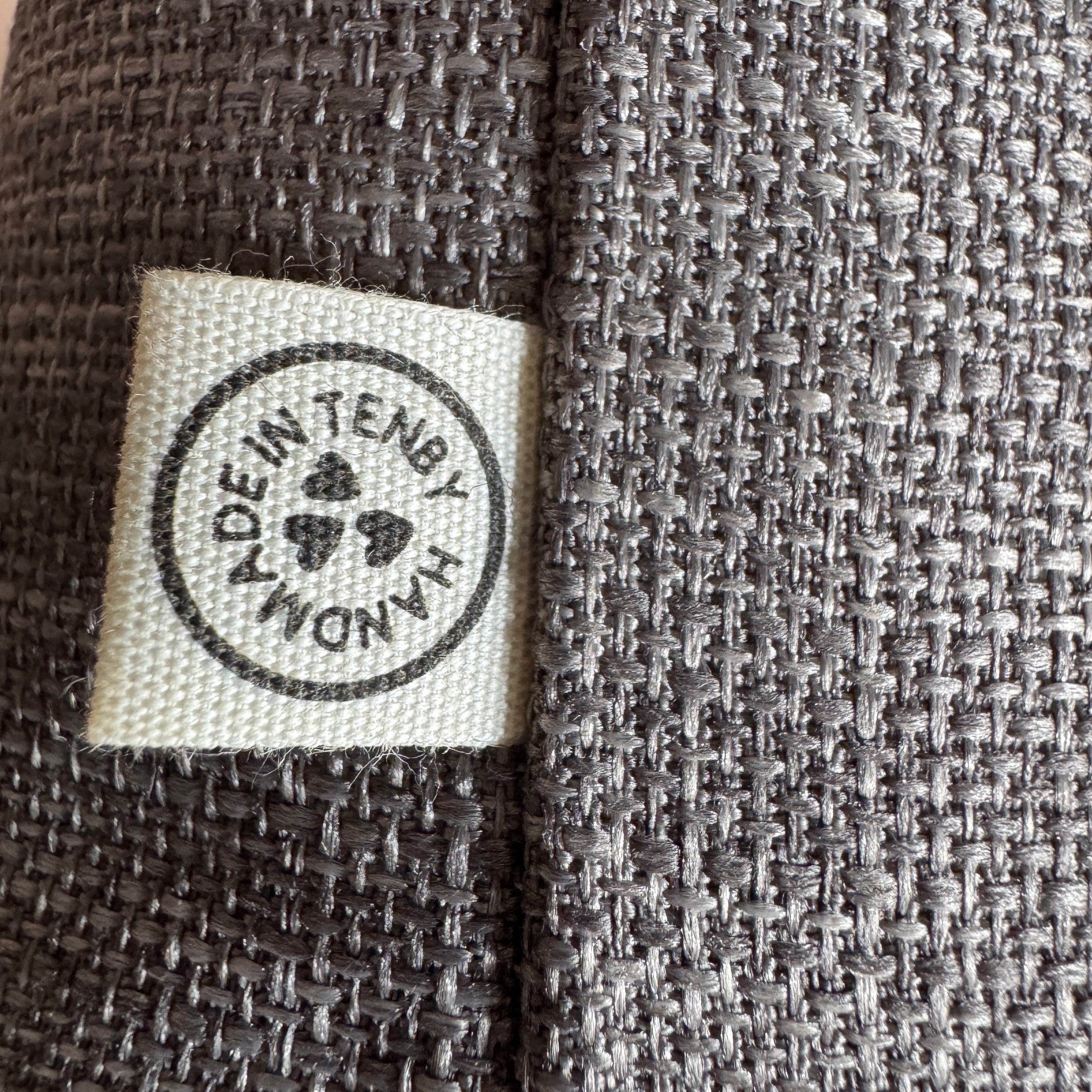 Close-up of a dark gray woven fabric with a square cream fabric label sewn onto the side. The label has a black circular stamp that reads 'MADE IN TENBY.