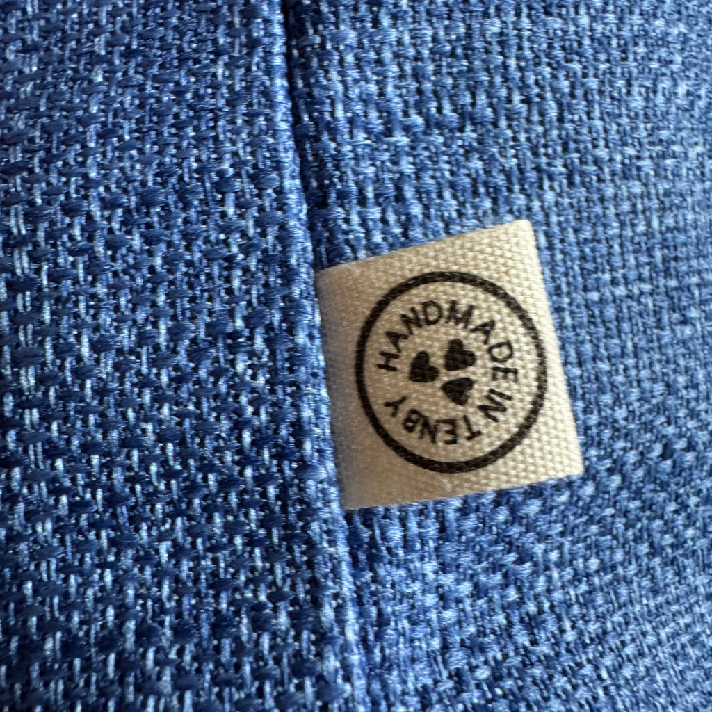Close-up of a textured blue fabric with a small beige fabric label sewn onto the side, featuring a circular black stamp that reads 'HANDMADE IN TENBY' with hearts