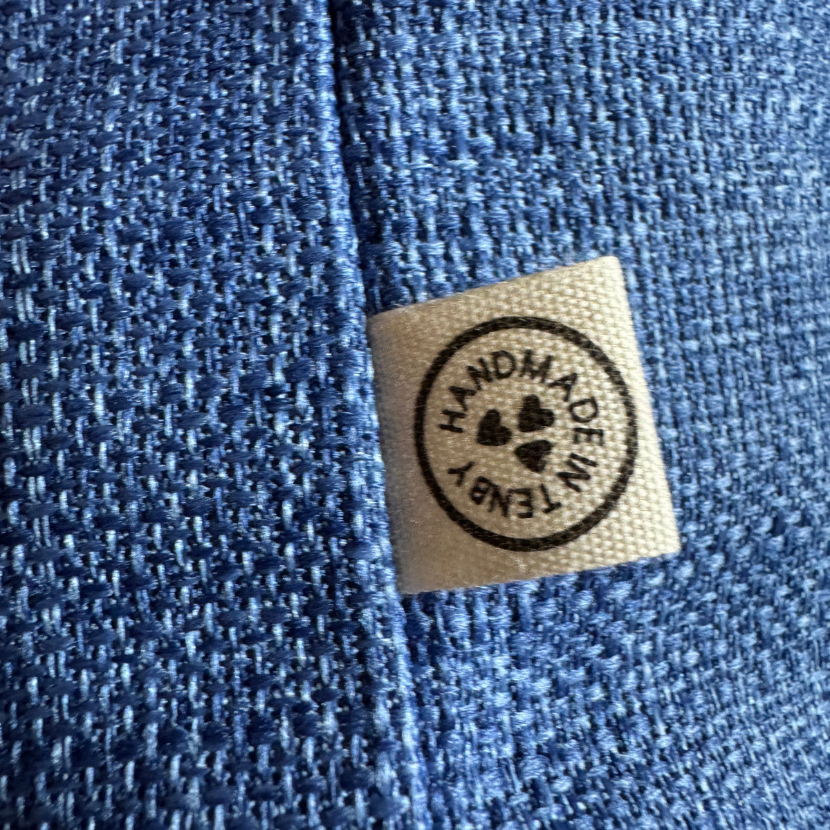 Close-up of a textured blue fabric with a small beige fabric label sewn onto the side, featuring a circular black stamp that reads 'HANDMADE IN TENBY' with hearts